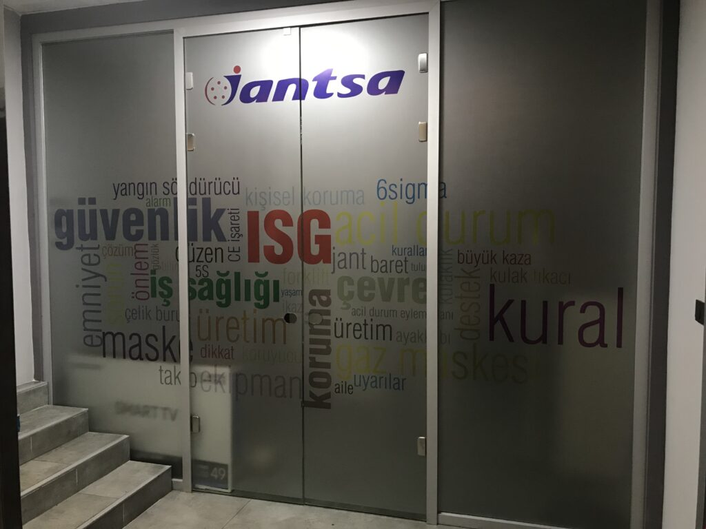Fabrika cam kapılarına uygulanan iş güvenliği ve iş sağlığı kelimelerinden oluşan dekoratif kumlu folyo çalışması